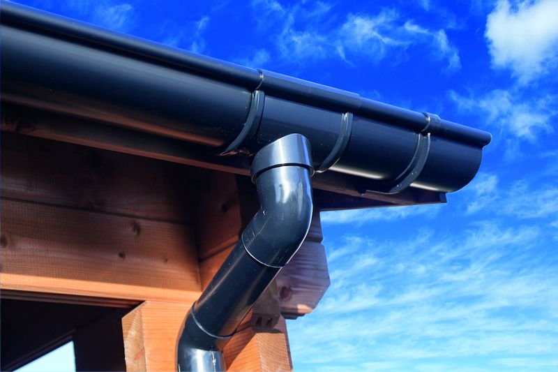 Gouttière et tuyau de descente en PVC noir fixés au toit d’un abri en bois contre un ciel bleu.